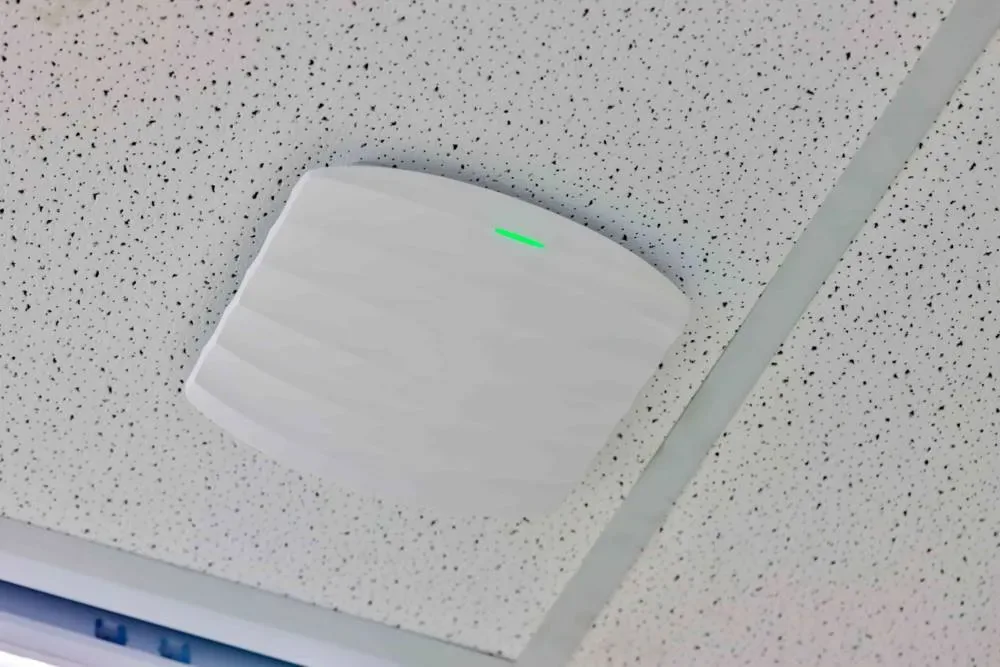 White ceiling-mounted wireless access point with a green status light on a drop ceiling tile