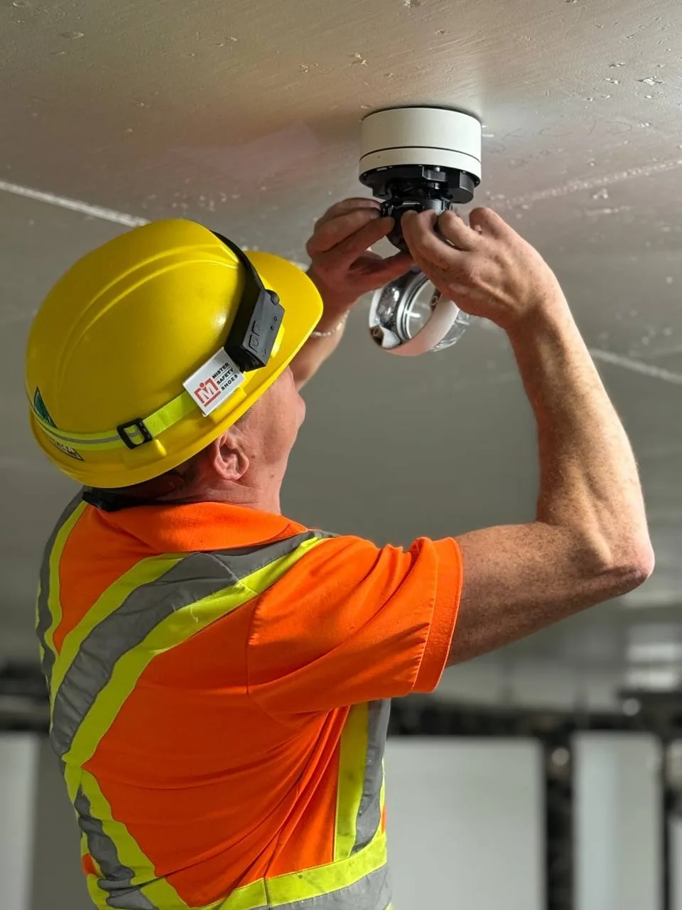 Technician in a yellow hard hat installing a dome camera on a concrete garage ceiling