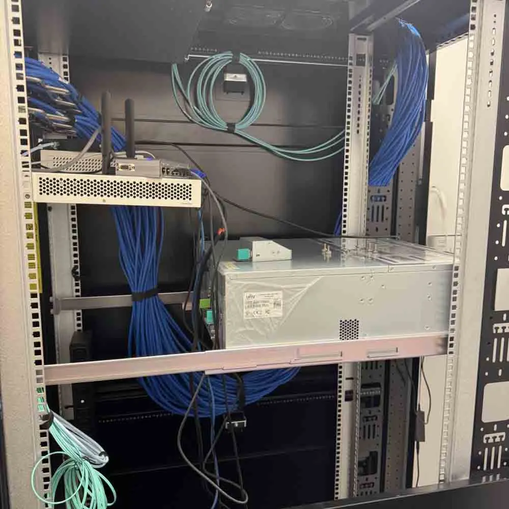 Structured cabling inside a network cabinet showing blue and green Ethernet cables, routers with antennas, and a Uniview power supply unit, organized for commercial network infrastructure.