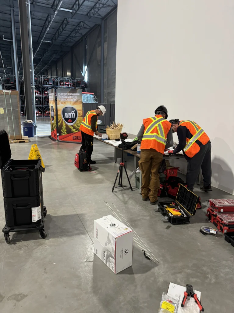 Technicians wearing high-visibility safety vests installing security and network equipment along a warehouse wall, with tools and cabling on the floor during a commercial surveillance system setup.