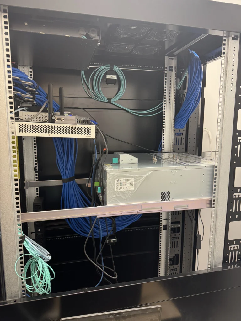 Open server rack cabinet showing organized blue Ethernet cables, fiber patch cords, network switch, and mounted equipment shelf, supporting commercial IT and security system infrastructure.
