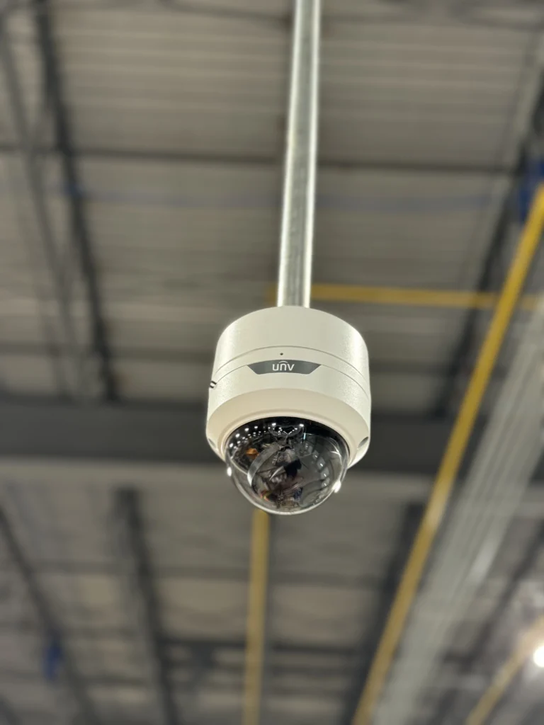 White dome security camera suspended from a high warehouse ceiling using a pendant mount, providing wide-area indoor surveillance coverage for industrial monitoring.
