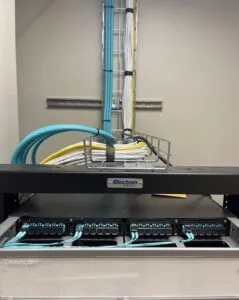 Wall-mounted network rack cabinet with organized blue Ethernet cables routed through cable management tray into patch panels and network switches, supporting commercial IT and security infrastructure.