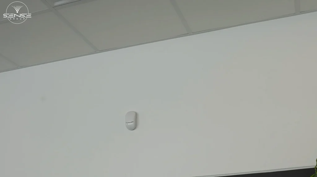 White motion detector sensor installed on an interior office wall near the ceiling, used as part of a commercial security alarm system to detect movement and unauthorized access.