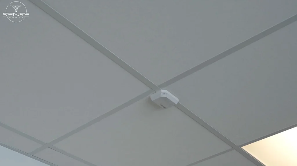 White wireless access point mounted on a drop ceiling tile grid in a commercial interior space, positioned at the intersection of ceiling panels for optimal WiFi signal distribution.