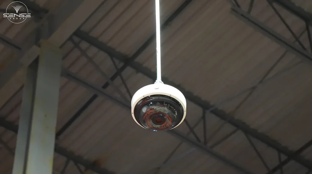 Dome CCTV security camera suspended from the ceiling in a large industrial warehouse, installed for wide-area video surveillance and high-ceiling monitoring coverage.