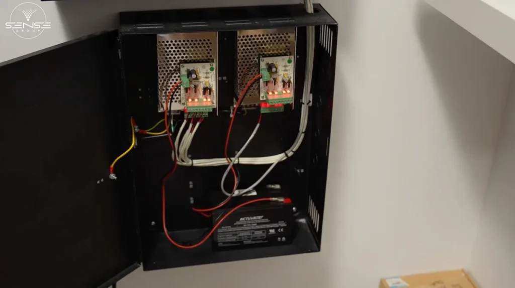 Open metal enclosure showing dual access control power supply modules with LED indicators, connected low-voltage wiring, and backup battery installed for secure door access system operation.