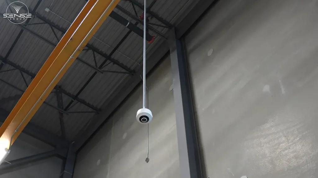 White dome security camera suspended from a high warehouse ceiling near a yellow overhead crane beam, installed for wide-area industrial video surveillance and facility monitoring.