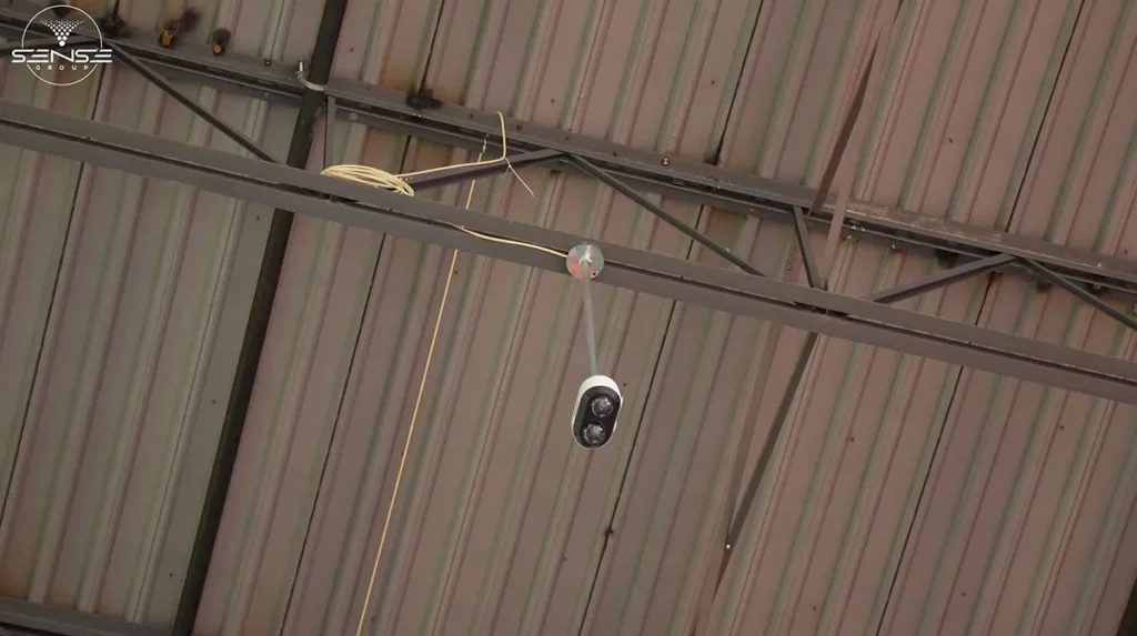 Bullet-style CCTV security camera mounted on a metal beam in a high industrial warehouse ceiling, positioned for long-range surveillance and wide-area monitoring coverage.