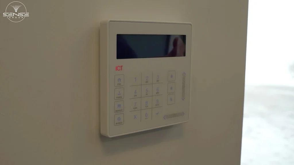 White ICT security alarm control panel mounted on an interior wall, featuring an LCD screen and numeric keypad for arming, disarming, and managing a home or office alarm system.