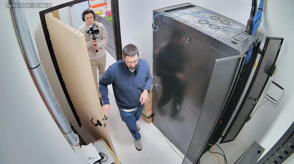 Overhead security camera view of a small server room with a tall network rack cabinet, structured Ethernet cabling, and two individuals entering the room under active video surveillance.