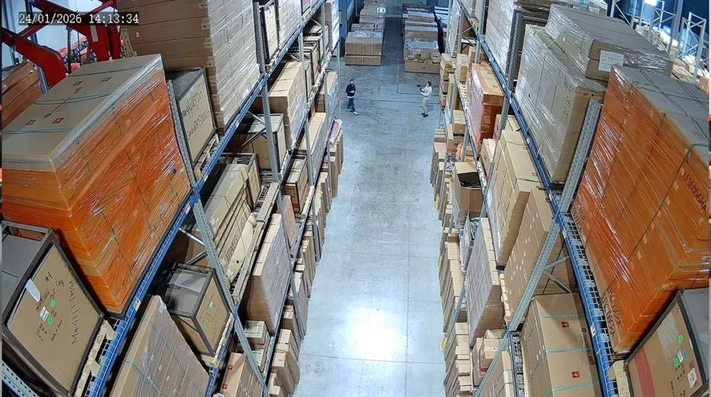 Overhead surveillance camera view of a warehouse aisle with tall storage racks filled with boxed inventory, showing two workers walking through the facility under active video monitoring.