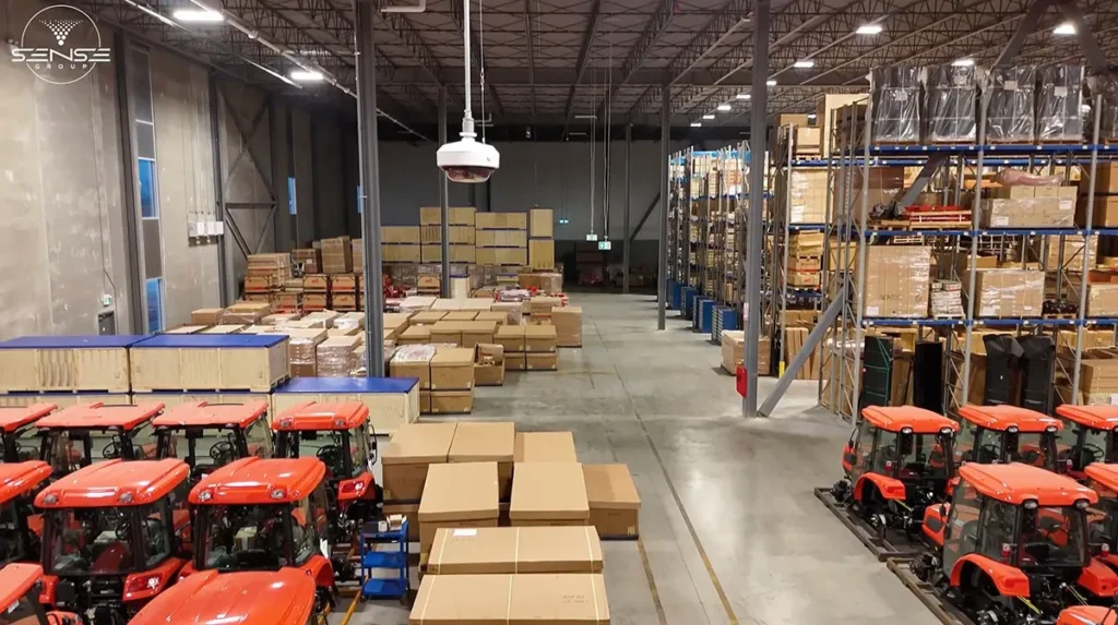 Large industrial warehouse interior with high ceilings, storage racks, boxed inventory, and red utility vehicles, monitored by a suspended dome security camera for wide-area video surveillance coverage.
