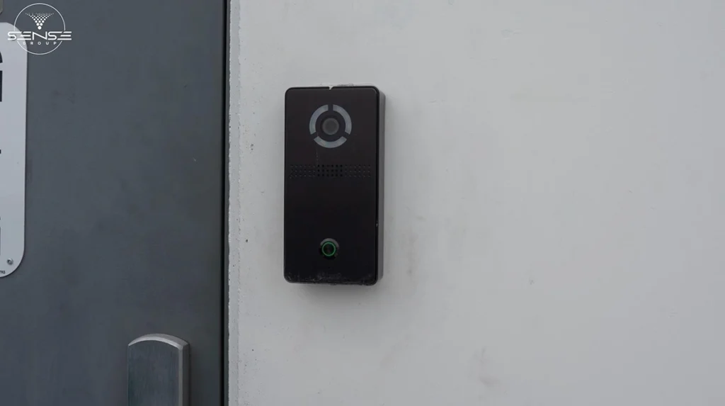 Black video doorbell intercom installed beside a commercial entrance door, featuring built-in camera, speaker grille, and illuminated call button for secure visitor access control.